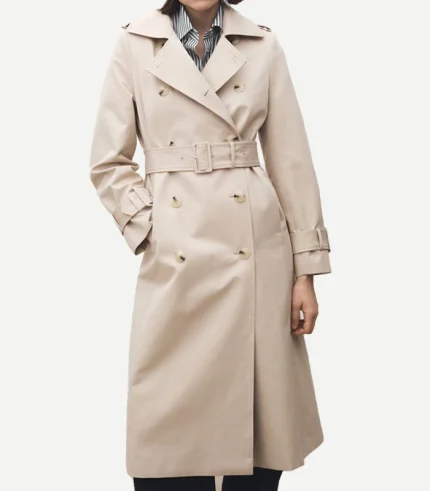 A person wearing a double-breasted beige cotton trench coat over a striped shirt. One hand is tucked into a side pocket, and the wide notched lapels are spread open. The coat is secured with a matching fabric belt with a rectangular buckle.