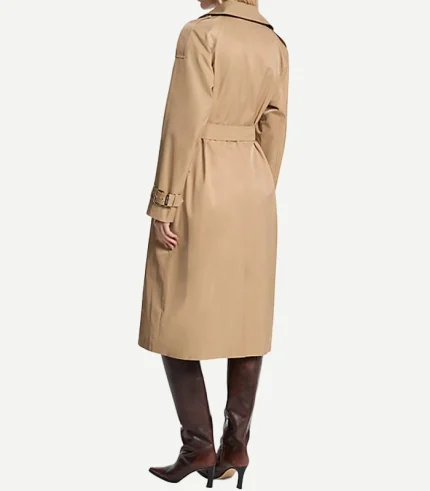 A woman seen from behind wearing a mid-length, tan cotton trench coat. The coat features a matching waist belt, structured epaulets on the shoulders, and adjustable buckled straps at the cuffs. She is also wearing knee-high, dark brown leather boots with a pointed toe and stiletto heel.