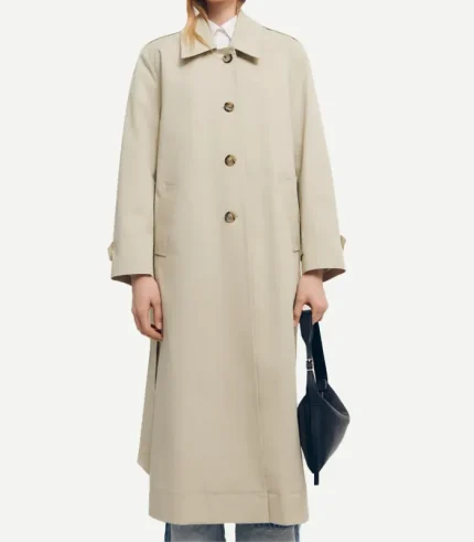 A front-facing view of a person wearing a long, beige cotton trench coat. The coat features a neat shirt-style collar, a single-breasted front with four large dark brown buttons, and subtle welt pockets. The silhouette is slightly oversized, and the person is holding a small black leather handbag.