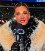 A medium shot of sports broadcaster Erin Andrews speaking into a microphone during an NFL broadcast. She is wearing a thick, high-collared fur coat in cream with black-tipped fur accents around the neck. She is wearing black leather gloves and gold hoop earrings.
