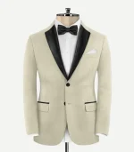 Front view of a slim-fit, light beige wool tuxedo jacket on a mannequin. It features contrasting black satin peak lapels, two black buttons, and jetted pockets with black satin trim. The jacket is styled with a white formal shirt and a black bow tie.