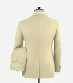 Back view of a men’s beige "Spring Latte" tuxedo jacket on a mannequin. The image shows a clean, tailored fit with a single center vent at the hem and a textured wool fabric finish. The matching trousers are shown draped over the arm of the mannequin, displaying the back pocket with a button closure and belt loops.