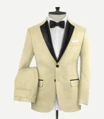 Front view of a men’s "Spring Latte" beige wool tuxedo suit on a mannequin. Features a slim-fit jacket with black satin peak lapels, a two-button closure with black fabric-covered buttons, and jetted pockets with black satin piping. The ensemble is styled with a white dress shirt, a black satin bow tie, and a white pocket square. Matching beige trousers are draped over the side.