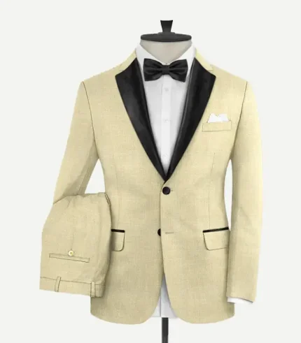 Front view of a men’s "Spring Latte" beige wool tuxedo suit on a mannequin. Features a slim-fit jacket with black satin peak lapels, a two-button closure with black fabric-covered buttons, and jetted pockets with black satin piping. The ensemble is styled with a white dress shirt, a black satin bow tie, and a white pocket square. Matching beige trousers are draped over the side.