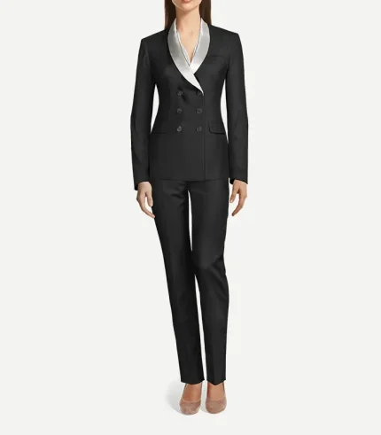 A full-length front view of a woman wearing a black, double-breasted tuxedo suit. The blazer features a slim fit, four black buttons, and a contrasting silver satin shawl lapel. The look is completed with matching black slim-fit trousers and pointed-toe heels.