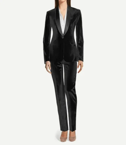 A woman wearing a black velvet tuxedo suit featuring a shawl lapel. The single-breasted jacket has a curved satin collar and a single button. It is styled with matching velvet trousers and neutral-toned pointed-toe shoes.