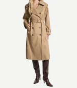 A front-facing view of a woman wearing a classic double-breasted tan cotton trench coat. The coat is styled with large notched lapels, dark tortoiseshell buttons, and a buckled belt cinched at the waist. She has one hand in a side pocket and is wearing the same knee-high, dark brown leather stiletto boots.
