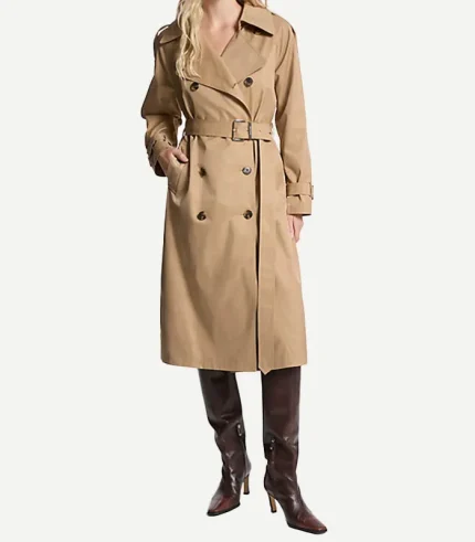 A front-facing view of a woman wearing a classic double-breasted tan cotton trench coat. The coat is styled with large notched lapels, dark tortoiseshell buttons, and a buckled belt cinched at the waist. She has one hand in a side pocket and is wearing the same knee-high, dark brown leather stiletto boots.