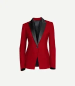 A front-facing close-up of a women’s red velvet tuxedo jacket. The design includes bold, black satin peak lapels, a single black button closure, and two horizontal jetted pockets at the waist. The interior is finished with a dark lining that is visible at the open neckline and cuffs.