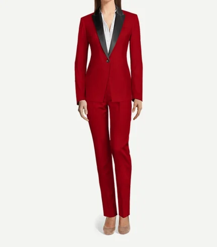 A full-length front view of a woman wearing a red velvet tuxedo. The single-button blazer features sharp, contrasting black satin peak lapels and is paired with matching slim-fit red velvet trousers. She is wearing a white collared shirt underneath and neutral-toned pointed-toe flats.