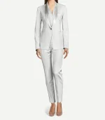 A woman modeling a tailored, monochromatic white velvet tuxedo. The outfit features a slim-fit blazer with a satin shawl lapel, a single gold button closure, and matching slim-leg trousers with a sharp front crease. She is wearing a white button-down shirt underneath and neutral-toned pointed-toe flats.