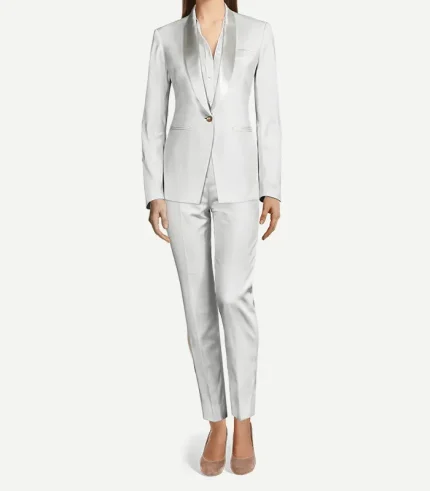 A woman modeling a tailored, monochromatic white velvet tuxedo. The outfit features a slim-fit blazer with a satin shawl lapel, a single gold button closure, and matching slim-leg trousers with a sharp front crease. She is wearing a white button-down shirt underneath and neutral-toned pointed-toe flats.