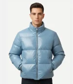 A male model wearing a metallic blue puffer jacket with his hands in his pockets. The jacket has a glossy finish, a full-zip front, and elasticated cuffs, presenting a modern, high-shine winter fashion look.