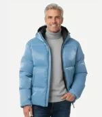 A man with grey hair smiling while wearing an open metallic light blue puffer jacket over a grey turtleneck and blue jeans. This version of the jacket includes an attached hood with a dark interior lining.