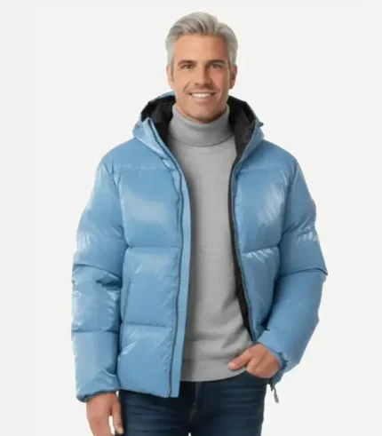 A man with grey hair smiling while wearing an open metallic light blue puffer jacket over a grey turtleneck and blue jeans. This version of the jacket includes an attached hood with a dark interior lining.