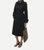A person is shown from the neck down wearing a Black Double Breasted Cotton Trench Coat. The coat is cinched at the waist with a matching buckled belt and features shoulder epaulets, adjustable buckled cuffs, and a wide collar. The person is carrying a large snakeskin-print tote bag and wearing black platform flip-flop sandals.