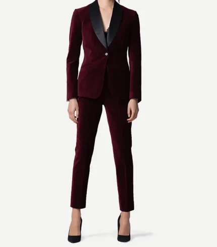 A woman wearing a tailored burgundy velvet tuxedo suit. The blazer features a deep black satin shawl lapel and a single-button closure, paired with matching slim-fit velvet trousers. She is wearing black pointed-toe heels against a plain white background.