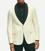 A front-facing view of a man wearing a classic-fit off-white dinner jacket featuring a prominent black contrast shawl lapel. The jacket has a single-button closure, flap pockets, and a welt chest pocket. He is styled with a black pleated tuxedo shirt, a black bowtie, and black trousers.