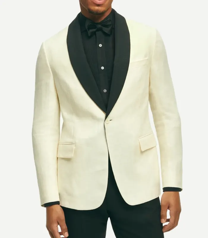 classic-fit-wool-white-dinner-jacket A front-facing view of a man wearing a classic-fit off-white dinner jacket featuring a prominent black contrast shawl lapel. The jacket has a single-button closure, flap pockets, and a welt chest pocket. He is styled with a black pleated tuxedo shirt, a black bowtie, and black trousers.