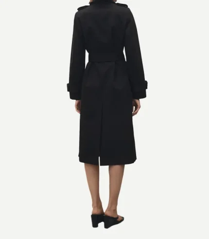 The back view of a person wearing the black cotton trench coat, emphasizing its tailored silhouette. The image shows the back of the buckled waist belt, the shoulder epaulets, and a central vent at the bottom hem for ease of movement. The person is wearing black heeled sandals.