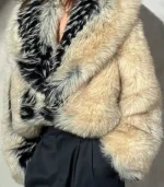 A detailed front view of the textured fur jacket. The image highlights the contrast between the soft cream-colored body and the thick, shaggy black and white fur lining the wide collar. The jacket is styled with high-waisted black trousers.