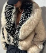 A close-up shot of a person wearing a voluminous, oversized fur jacket. The jacket features a cream-colored base with distinct vertical stripes of black and white speckled fur along the lapel and sleeves. The person is wearing a black baseball cap that partially obscures their face.