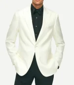 A front-facing view of a man wearing an off-white, slim-fit dinner jacket with notch lapels and a single-button closure. He is styled with a black pleated tuxedo shirt, a black bowtie, and black dress trousers. His hands are placed in the jacket's flap pockets.