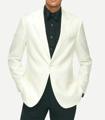 A front-facing view of a man wearing an off-white, slim-fit dinner jacket with notch lapels and a single-button closure. He is styled with a black pleated tuxedo shirt, a black bowtie, and black dress trousers. His hands are placed in the jacket's flap pockets.