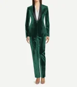 A full-length view of a woman wearing a tailored emerald green velvet tuxedo. She is styled with a crisp white button-down shirt underneath and neutral-toned pointed-toe heels. The suit features a black satin shawl lapel and straight-leg matching velvet trousers.