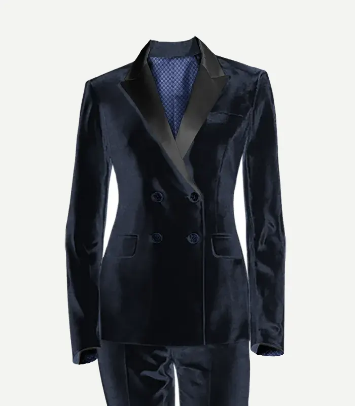 A studio product photograph of a women's navy blue velvet double-breasted tuxedo jacket. The jacket features sharp black satin peak lapels, four decorative front buttons, and a visible blue patterned lining at the neck. It is shown with matching navy velvet trousers against a clean white background.