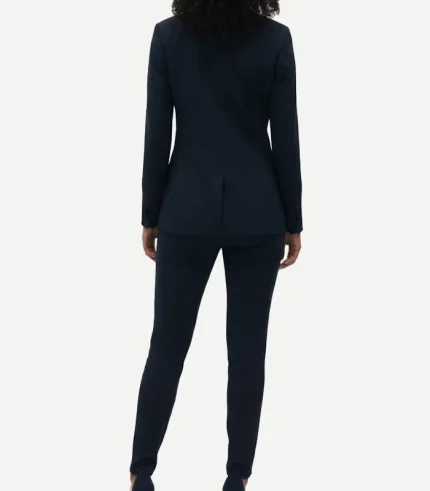 Rear view of a woman in a navy blue tuxedo blazer showing a clean shoulder line and center vent, paired with matching slim-cut formal pants.